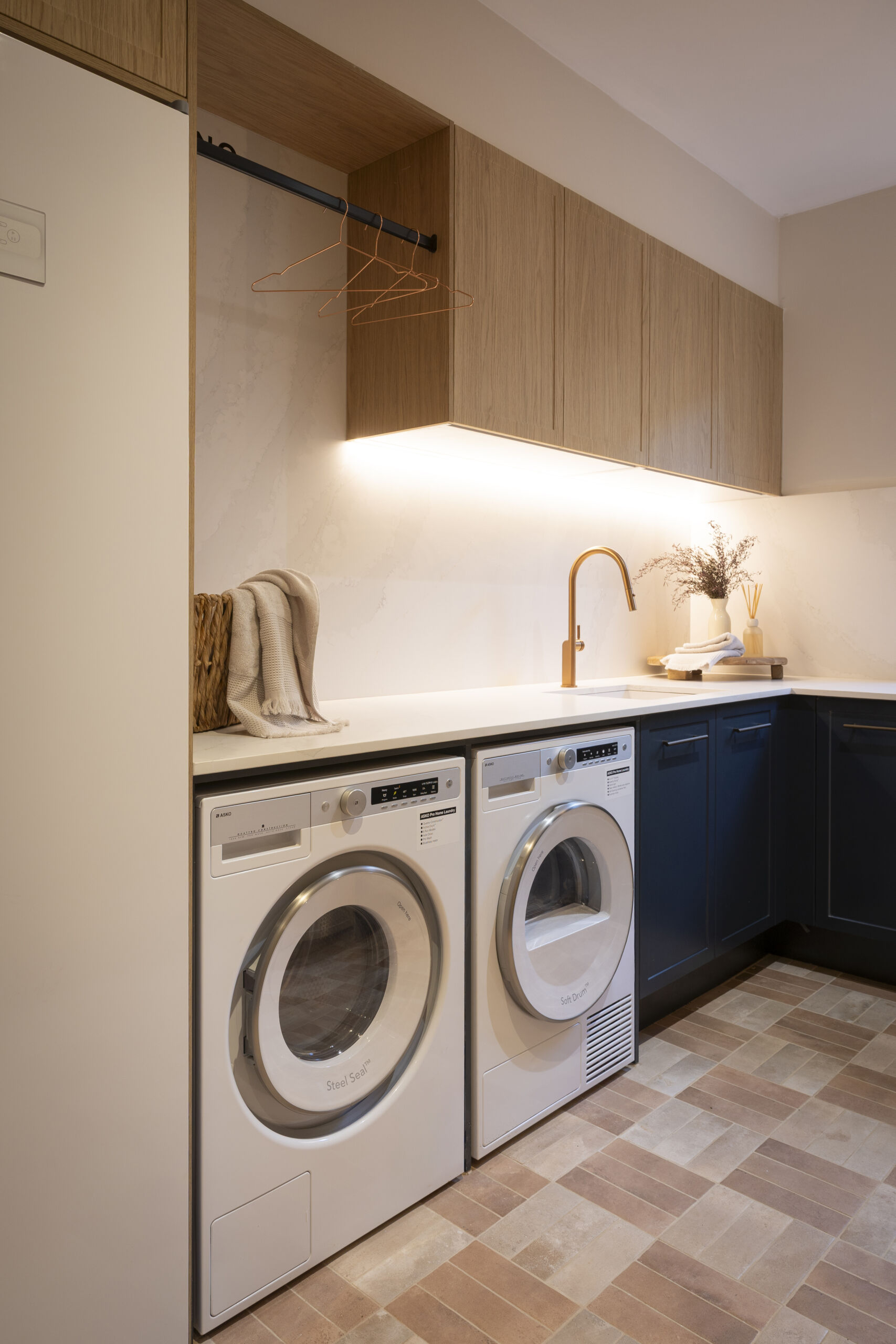 H4 SONNY & ALICIA Laundry MA261 Laundry with hanging rail under timber-look wall cabinets, navy base cabinetry and side-by-side washer and dryer