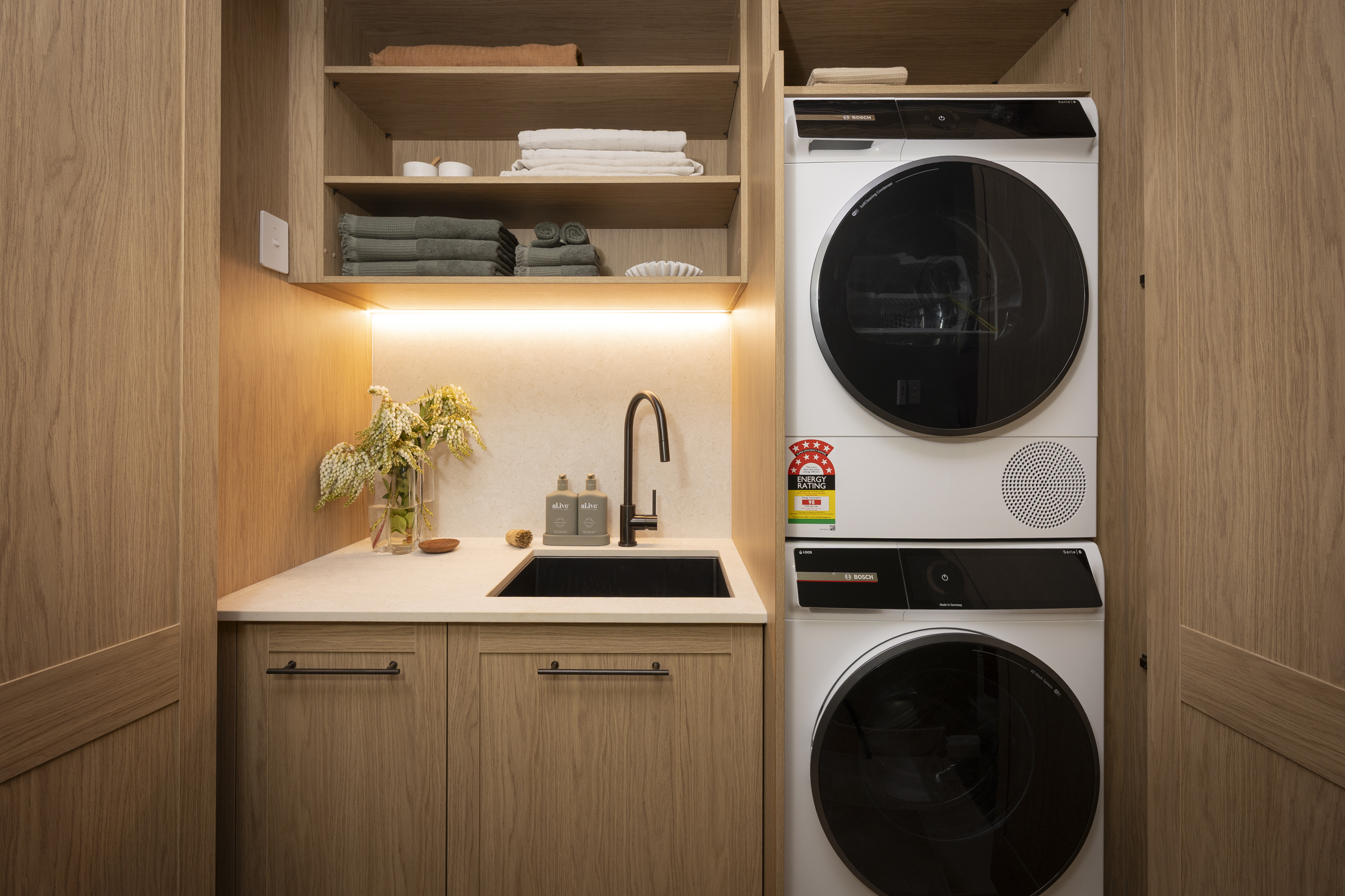 H5 ROBBIE & MAT Laundry MA_FM107 Small laundry with stacked washer and dryer, timber-look cabinetry, open shelving and a compact sink