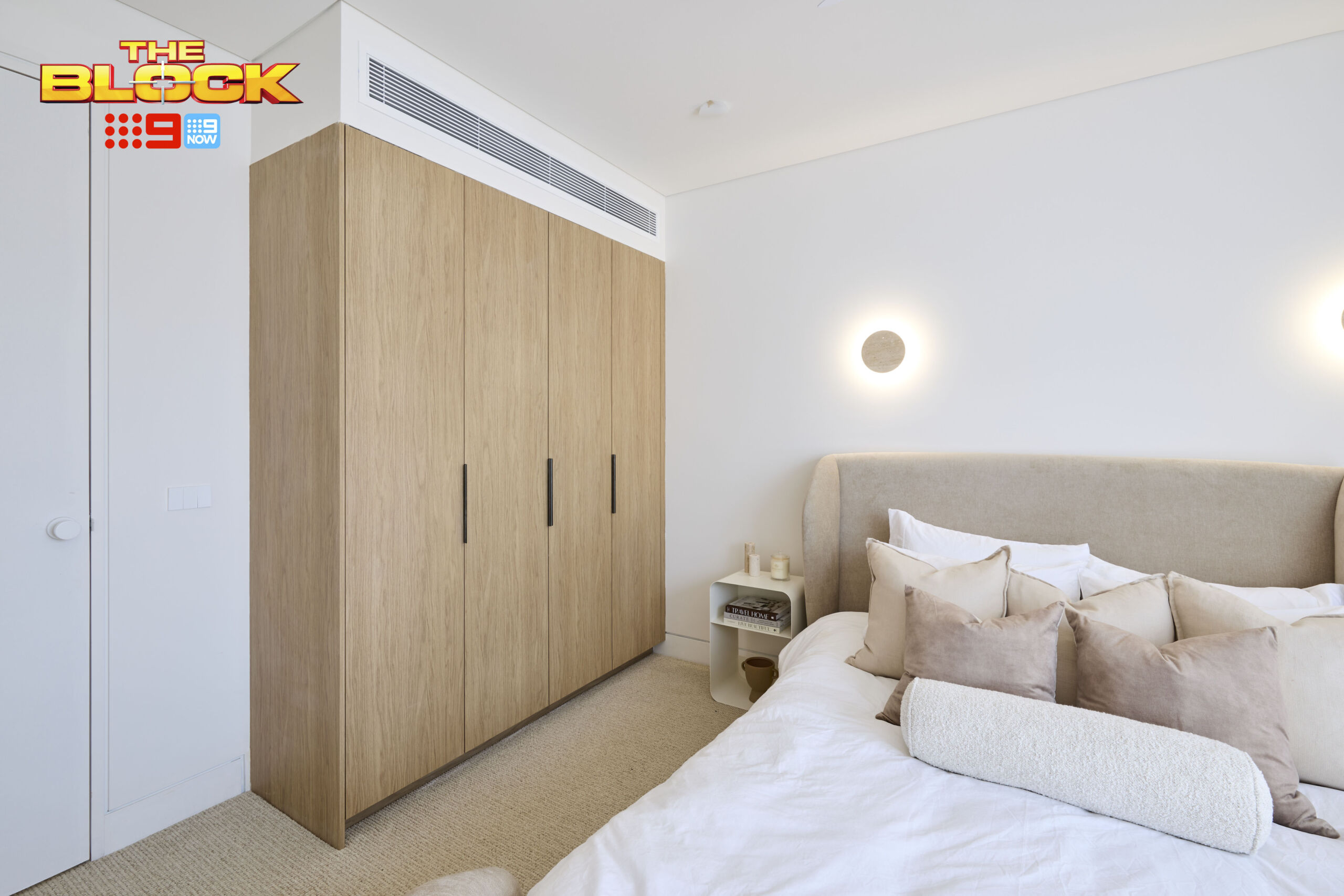 Built-in timber-look wardrobe with black handles in a contemporary guest bedroom featuring a soft upholstered bedhead and neutral styling.
