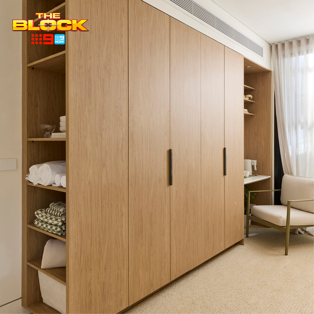 Freedom Wardrobes built-in wardrobe on The Block 2026, timber-toned cabinetry with open shelving to one side holding towels and linen, beside a curtained window and armchair.